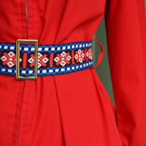 Puede incluir: Un vestido rojo con un cintur&oacute;n estampado en azul, blanco y rojo. El vestido tiene una cremallera en la parte delantera y mangas largas.
