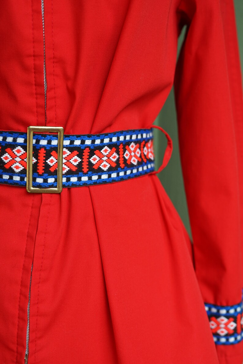 Puede incluir: Un vestido rojo con un cintur&oacute;n estampado en azul, blanco y rojo. El vestido tiene una cremallera en la parte delantera y mangas largas.
