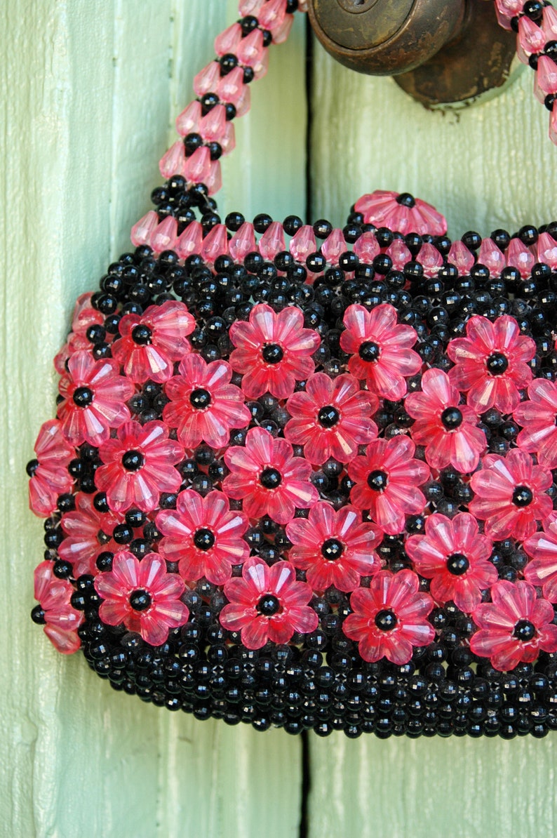May include: A pink and black beaded handbag with a floral design. The bag is made of small, round beads that are strung together to create a pattern of flowers.