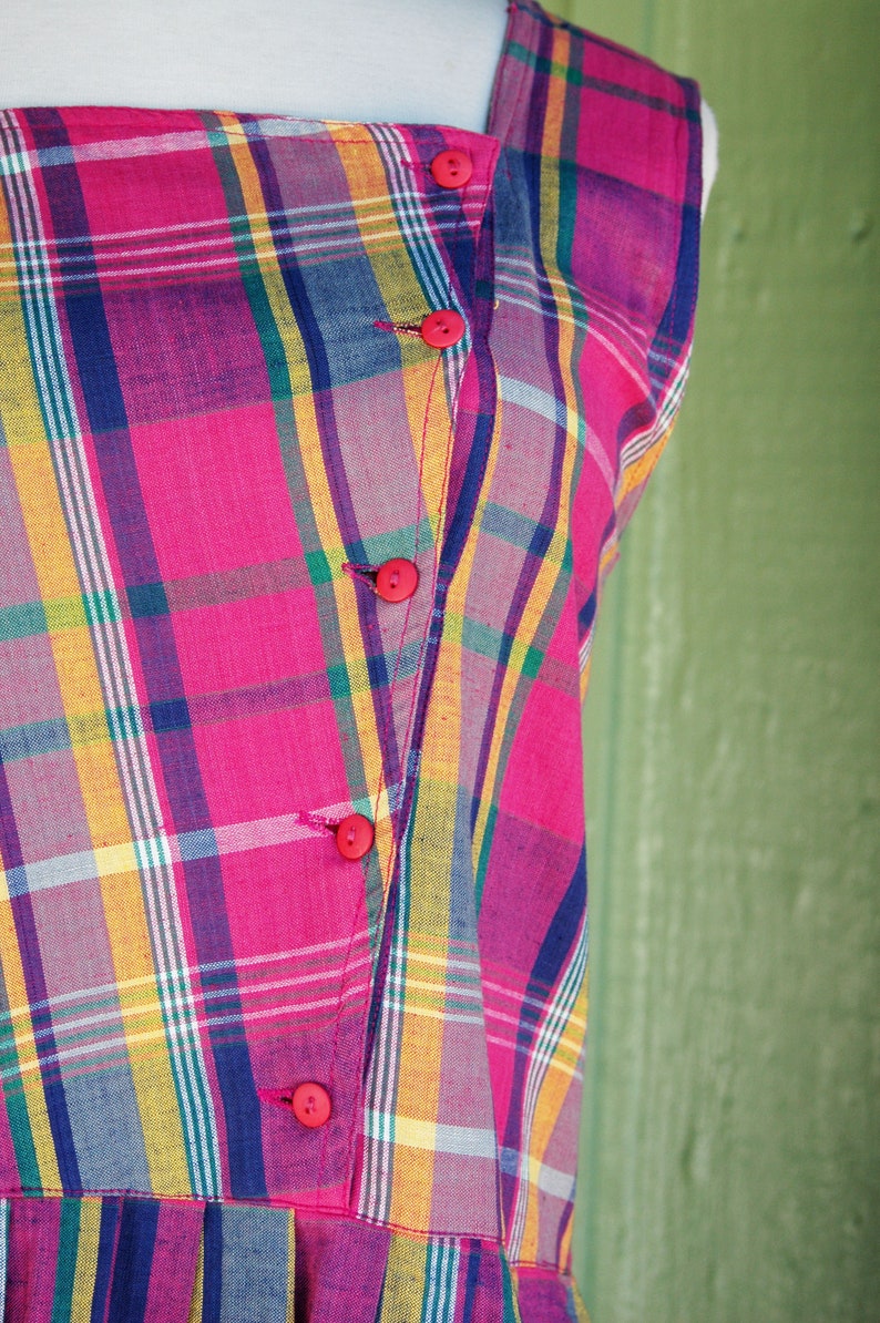 Pu&ograve; includere: Un abito senza maniche con un motivo a quadri rosa, giallo, blu e verde. L'abito ha una chiusura con bottoni sul davanti e una gonna a pieghe.