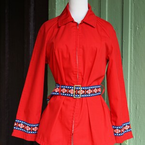 Puede incluir: Una chaqueta roja con cremallera con un cintur&oacute;n de estampado geom&eacute;trico y ribetes azules y blancos en las mangas.