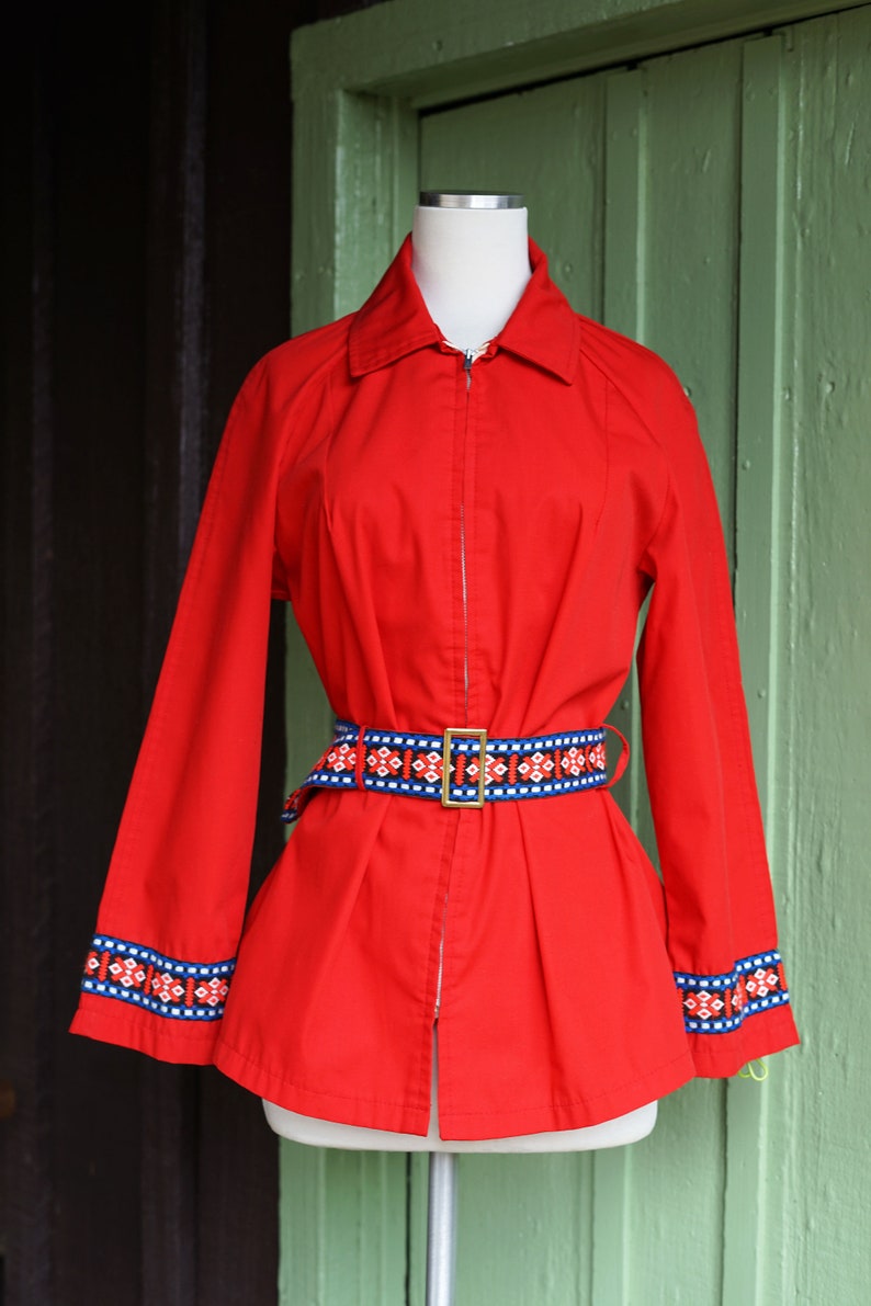 Puede incluir: Una chaqueta roja con cremallera con un cintur&oacute;n de estampado geom&eacute;trico y ribetes azules y blancos en las mangas.