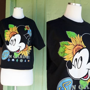 以下が含まれることがあります： ミッキーマウスとヒマワリが描かれた黒いクロップドTシャツ。ミッキーマウスの下に「Sun Garden!」の文字がプリントされています。