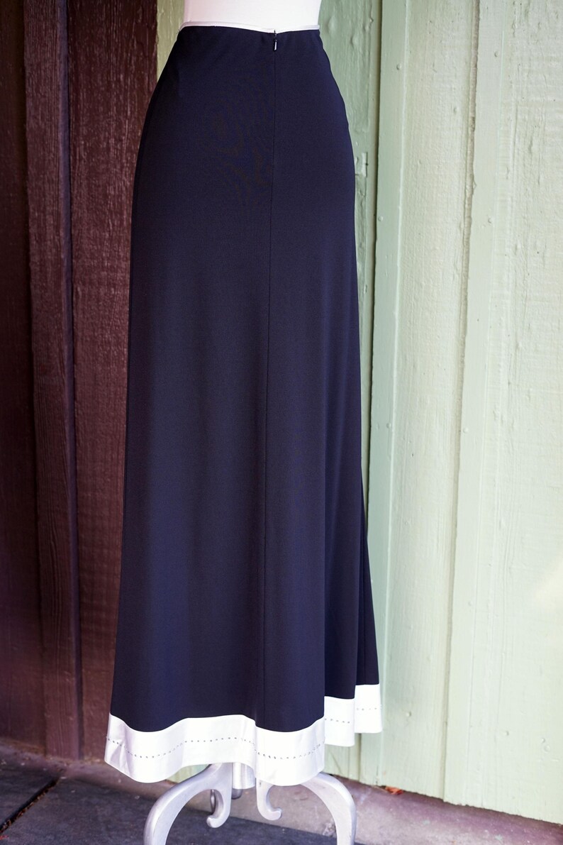 May include: A navy blue maxi skirt with a white hemline. The skirt has a simple, elegant design, with a straight silhouette and a contrasting white border at the bottom. The white border has a dotted pattern.
