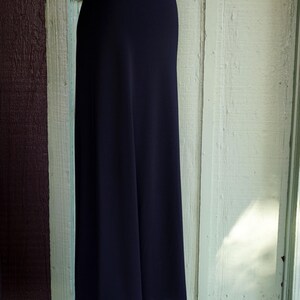 May include: A black, floor-length skirt with a white hem featuring a row of small, evenly spaced holes. The skirt is displayed on a mannequin against a backdrop of weathered wooden panels.