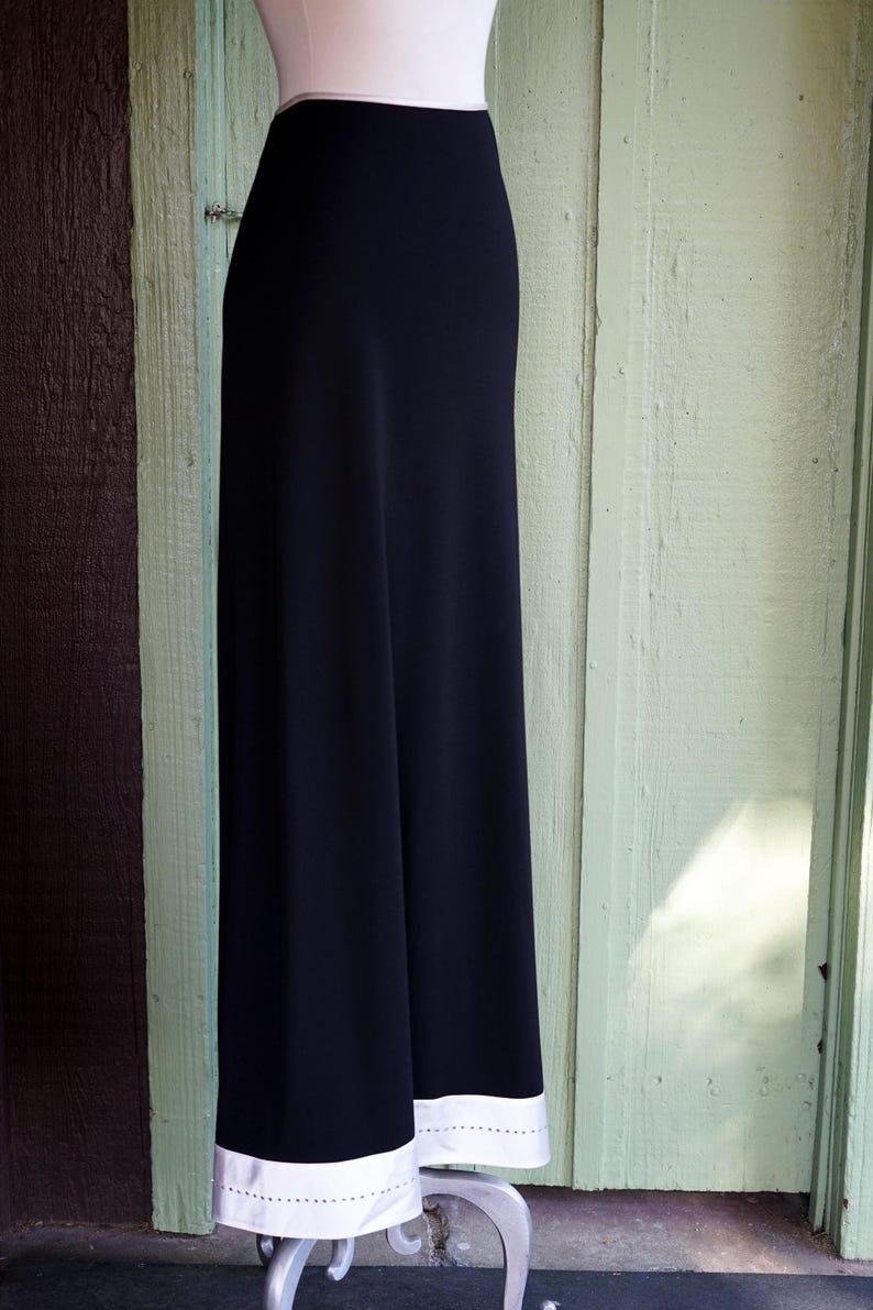 May include: A black, floor-length skirt with a white hem featuring a row of small, evenly spaced holes. The skirt is displayed on a mannequin against a backdrop of weathered wooden panels.