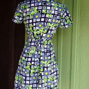 May include: A vintage dress with a floral pattern. The dress features a navy blue background with a white grid design, and is adorned with blue and yellow flowers and green leaves. The dress has short sleeves and a tie waist.
