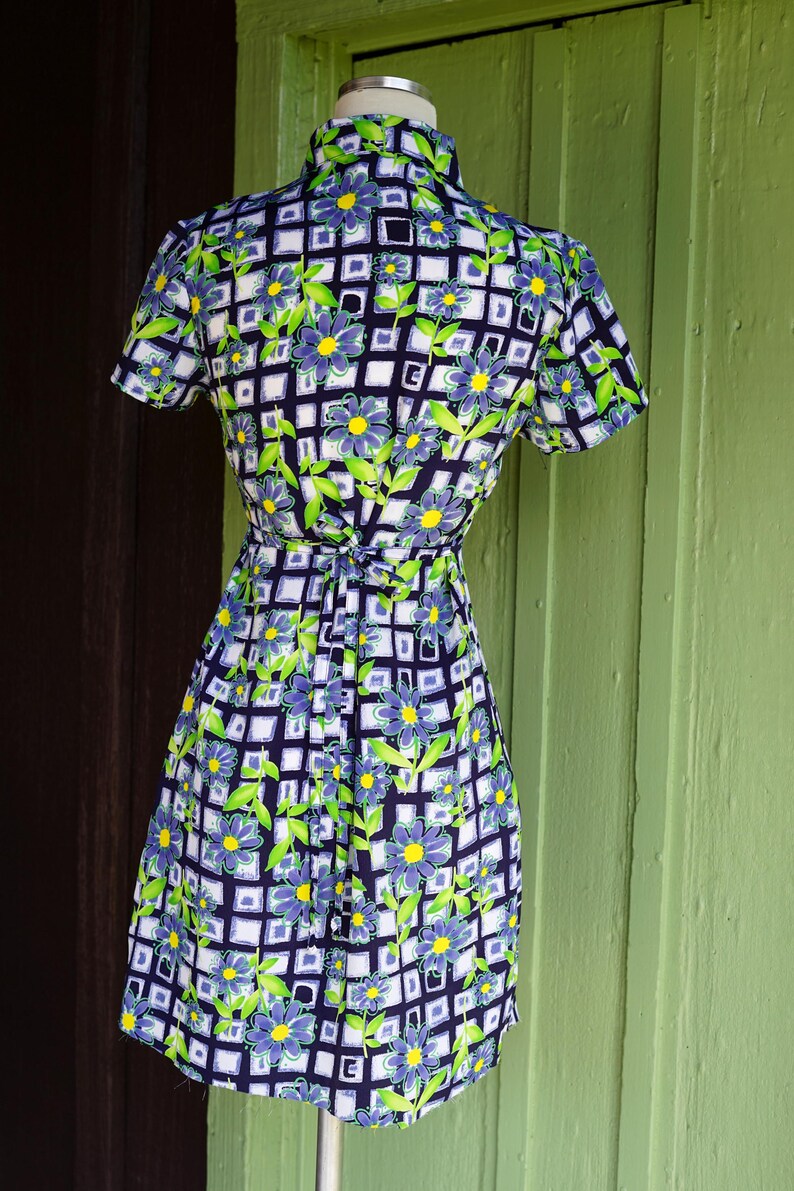 May include: A vintage dress with a floral pattern. The dress features a navy blue background with a white grid design, and is adorned with blue and yellow flowers and green leaves. The dress has short sleeves and a tie waist.