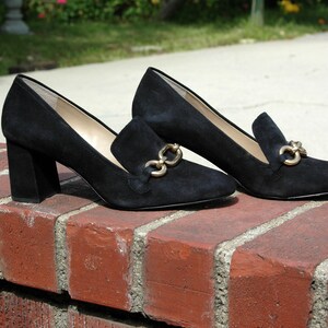 May include: A pair of black suede loafers with gold-tone chain accents. The shoes have a block heel and a classic slip-on design, perfect for a sophisticated look. The shoes are displayed on a red brick surface.