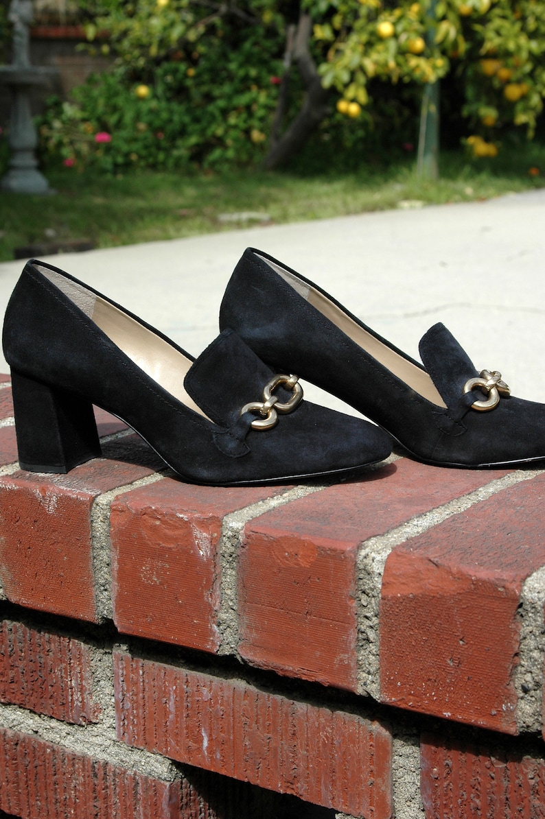 May include: A pair of black suede loafers with gold-tone chain accents. The shoes have a block heel and a classic slip-on design, perfect for a sophisticated look. The shoes are displayed on a red brick surface.