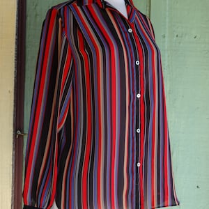 May include: A long-sleeved, striped button-down shirt in black, red, blue, and beige. The shirt is made of a sheer fabric and has a relaxed fit.
