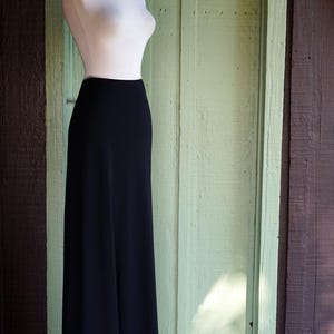May include: A black, floor-length skirt with a white trim and a mannequin torso. The skirt is displayed on a silver metal stand against a light green wooden door and a brown wooden wall.