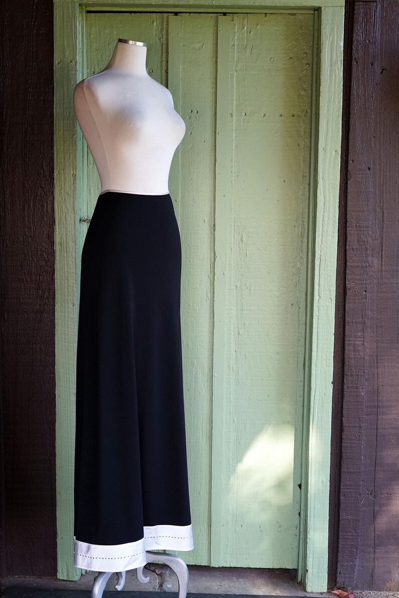 May include: A black, floor-length skirt with a white trim and a mannequin torso. The skirt is displayed on a silver metal stand against a light green wooden door and a brown wooden wall.