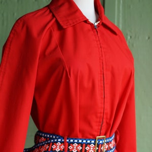 Puede incluir: Una chaqueta roja con cremallera y un cintur&oacute;n con estampado geom&eacute;trico de colores. La chaqueta tiene cuello y mangas largas.