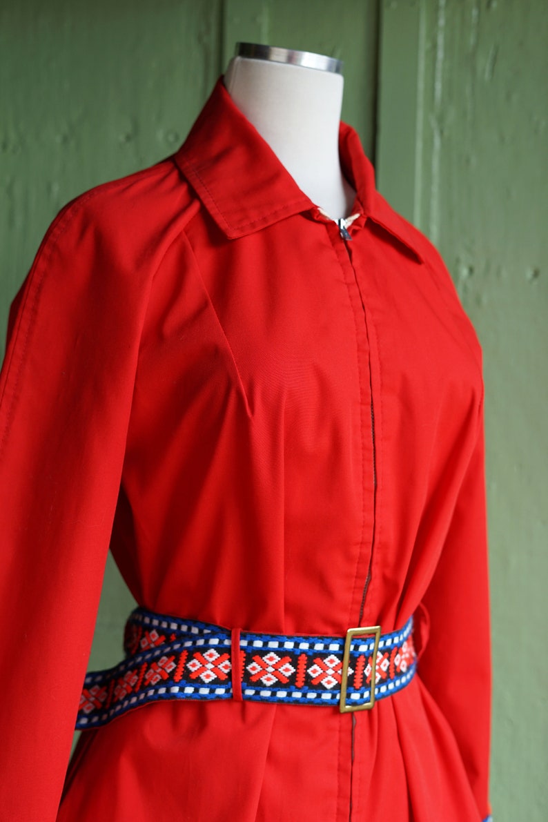 Puede incluir: Una chaqueta roja con cremallera y un cintur&oacute;n con estampado geom&eacute;trico de colores. La chaqueta tiene cuello y mangas largas.