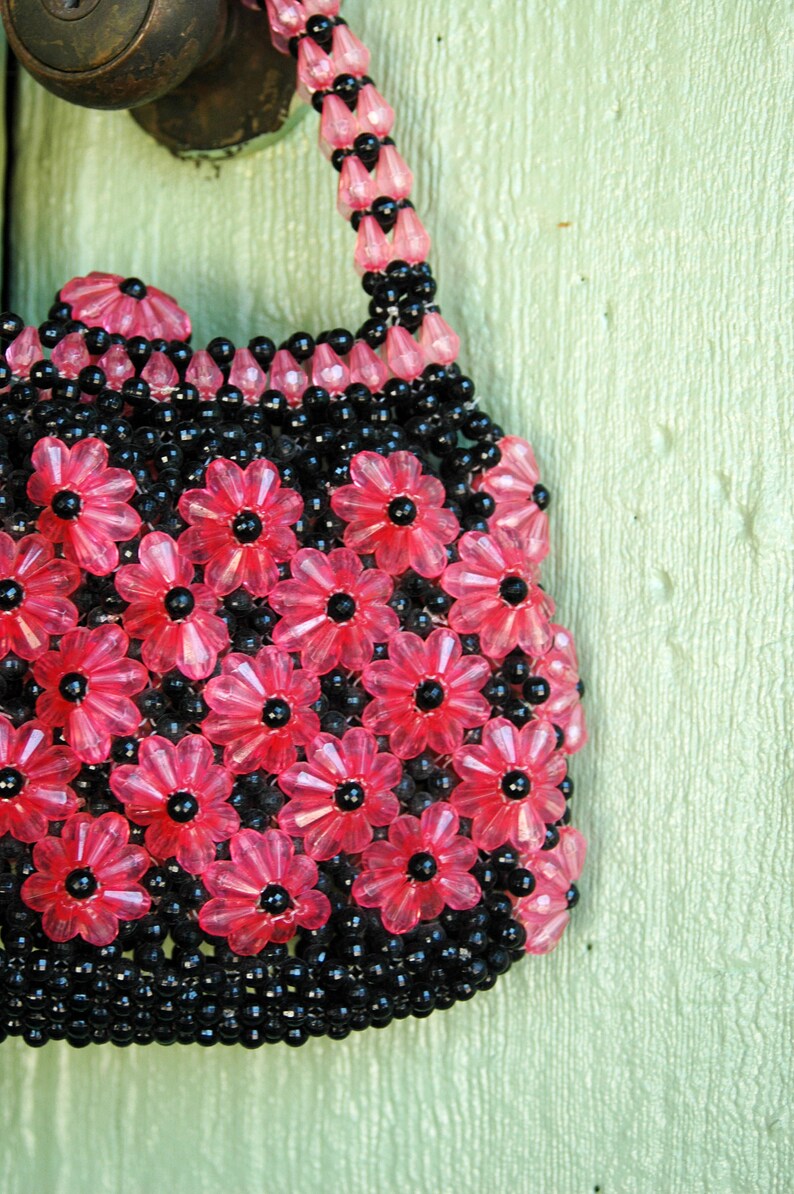 May include: A pink and black beaded purse with a flower design. The purse is hanging on a light green door.