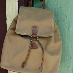 May include: A tan canvas backpack with brown leather straps and a metal ring. The backpack is hanging on a door handle.