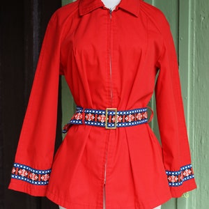 Puede incluir: Una chaqueta roja con cierre de cremallera y un cintur&oacute;n con estampado geom&eacute;trico. La chaqueta tiene ribetes azules y blancos en las mangas.