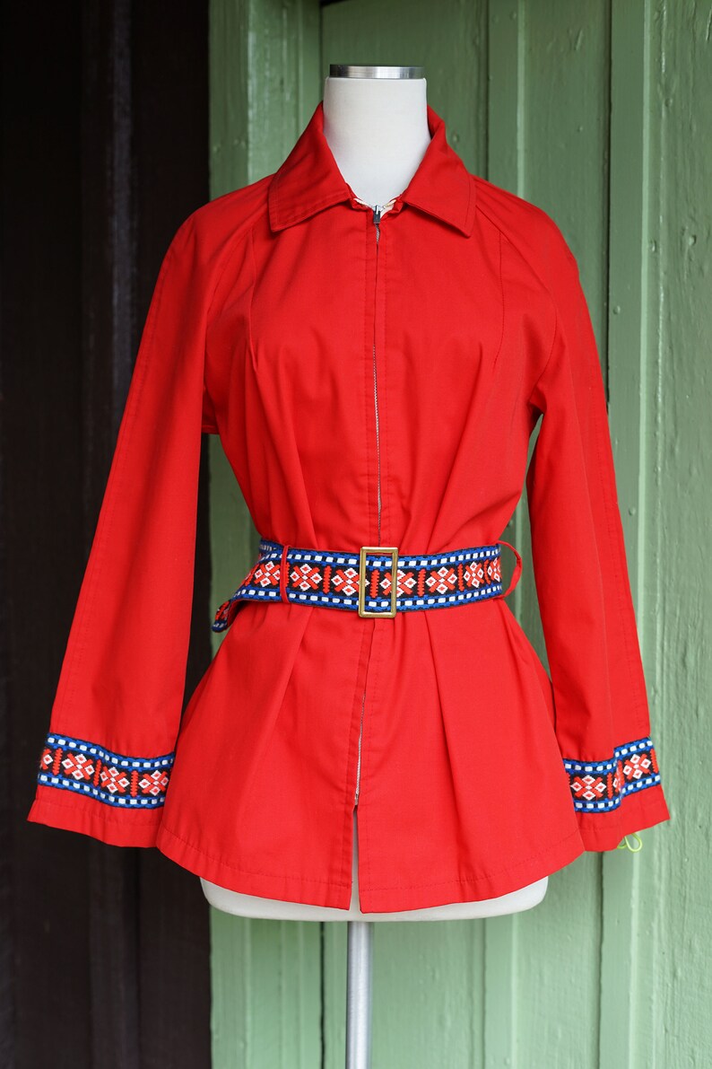 Puede incluir: Una chaqueta roja con cierre de cremallera y un cintur&oacute;n con estampado geom&eacute;trico. La chaqueta tiene ribetes azules y blancos en las mangas.