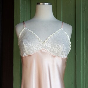 May include: A pink satin slip with a white lace trim. The slip is on a white mannequin and is displayed against a green background.