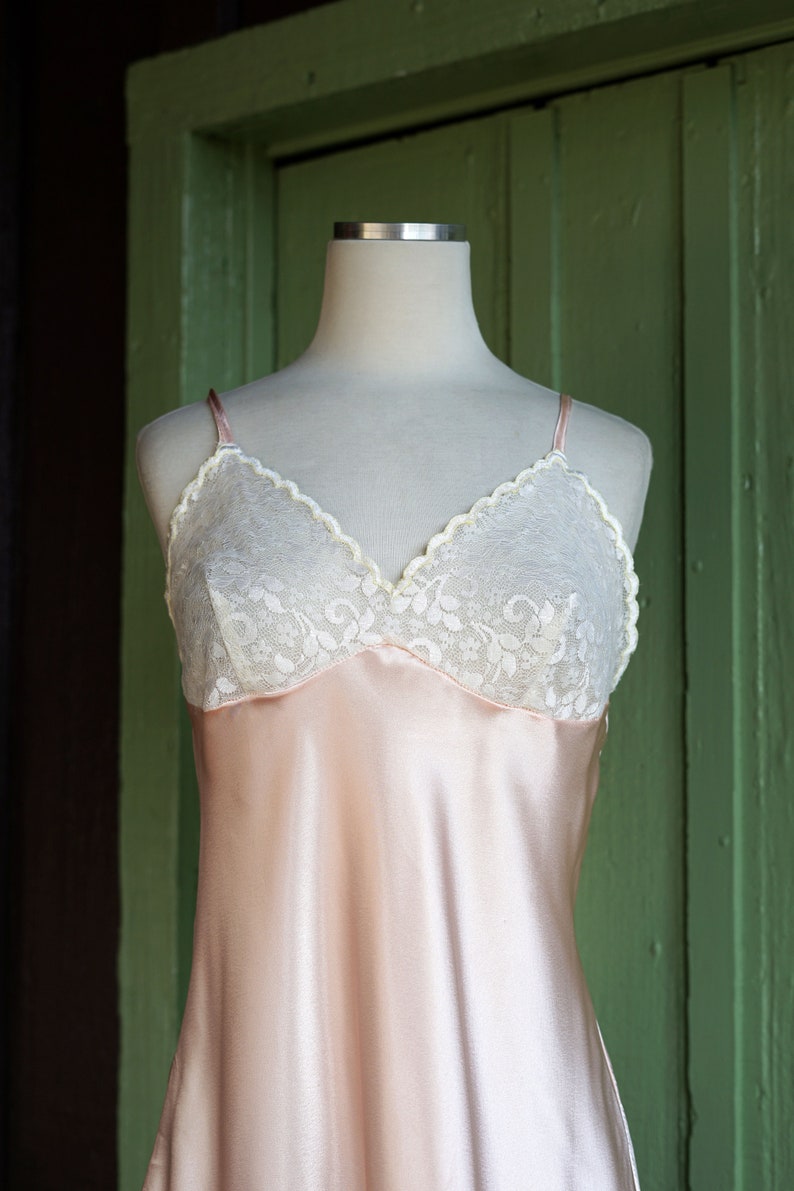 May include: A pink satin slip with a white lace trim. The slip is on a white mannequin and is displayed against a green background.