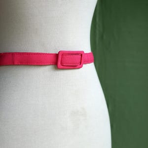 K&ouml;nnte beinhalten: Ein rosa Stoffg&uuml;rtel mit einer rechteckigen Schnalle. Der G&uuml;rtel befindet sich an einem wei&szlig;en Mannequin.
