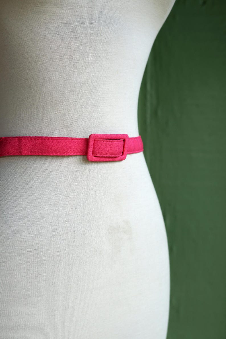 K&ouml;nnte beinhalten: Ein rosa Stoffg&uuml;rtel mit einer rechteckigen Schnalle. Der G&uuml;rtel befindet sich an einem wei&szlig;en Mannequin.