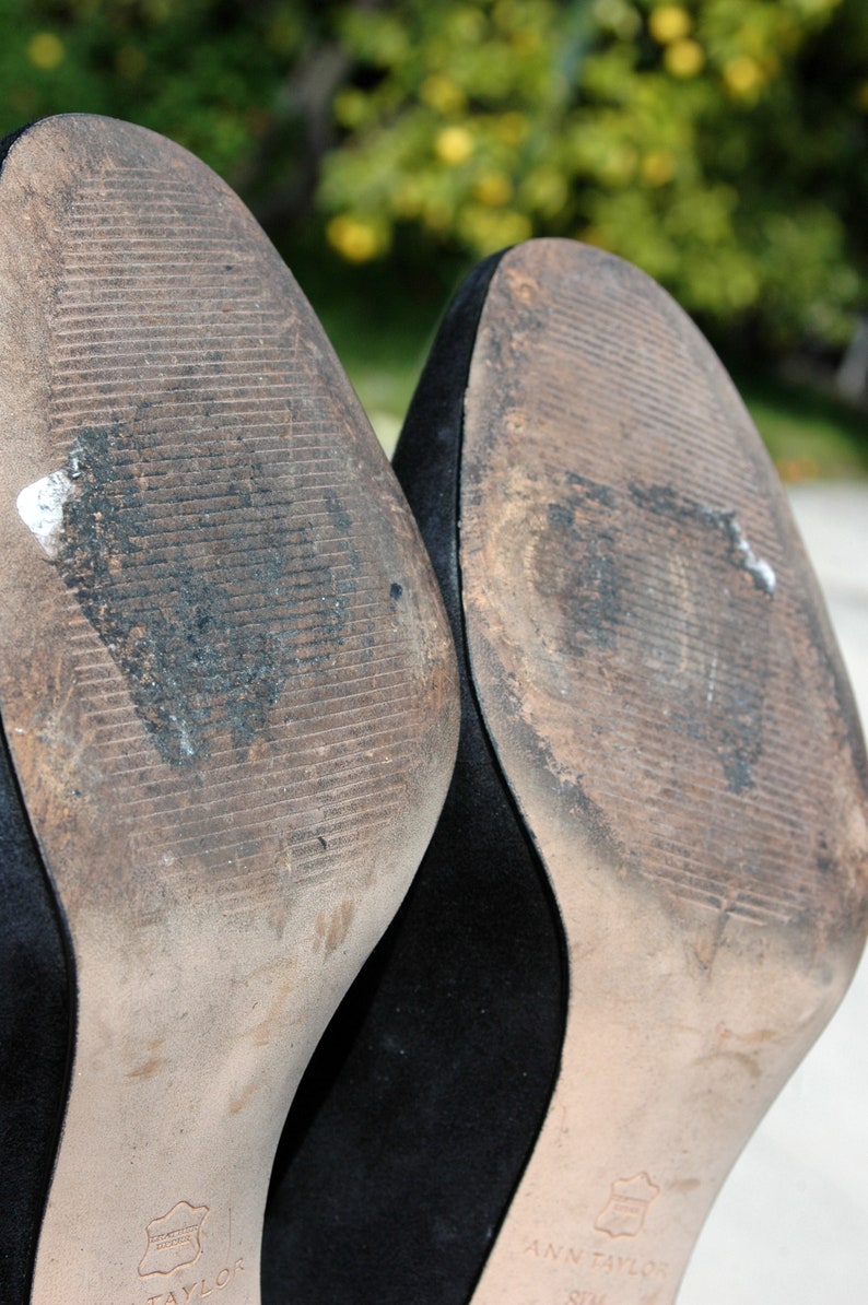 May include: Close-up of the soles of two black high-heeled shoes. The soles are worn and show signs of use, with dirt and wear patterns visible. The brand name "ANN TAYLOR" is visible on the sole.