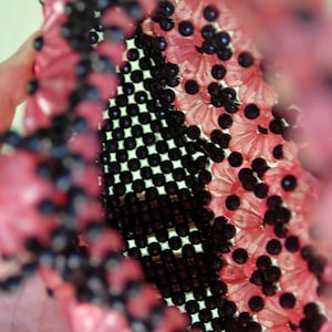 May include: A close-up of a pink and black beaded purse. The purse is made of small, round beads that are woven together in a pattern. The beads are pink and black, and the purse has a unique, textured look.