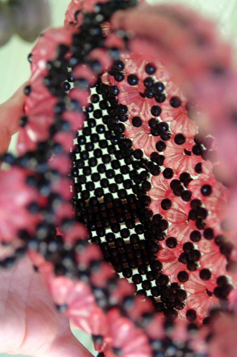 May include: A close-up of a pink and black beaded purse. The purse is made of small, round beads that are woven together in a pattern. The beads are pink and black, and the purse has a unique, textured look.