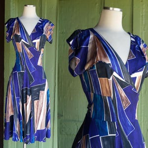 May include: A vintage blue dress with a geometric pattern. The dress features a V-neckline, short sleeves, and a wrap-style design. The pattern includes shades of blue, brown, black, and white. The dress is displayed on a mannequin.