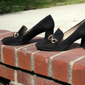 May include: A pair of black suede heeled loafers with gold-tone hardware. The shoes have a classic design with a block heel and a slip-on style. The loafers are displayed on a red brick wall.