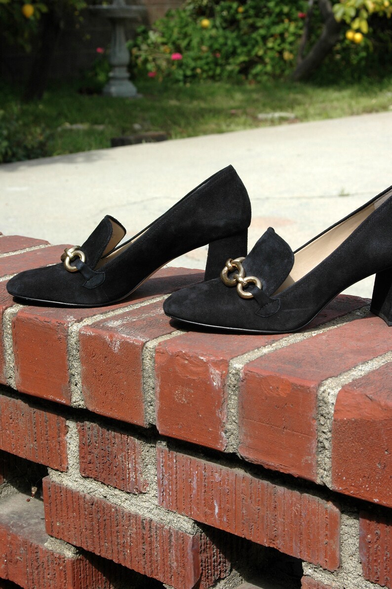 May include: A pair of black suede heeled loafers with gold-tone hardware. The shoes have a classic design with a block heel and a slip-on style. The loafers are displayed on a red brick wall.