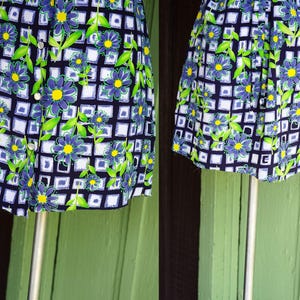 May include: A vintage floral print dress featuring a navy blue background with a white grid pattern. The dress is adorned with blue flowers with yellow centers and green leaves. It has a button-up front and is displayed on a mannequin.