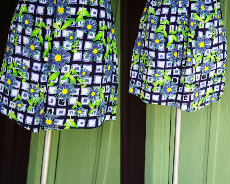 May include: A vintage floral print dress featuring a navy blue background with a white grid pattern. The dress is adorned with blue flowers with yellow centers and green leaves. It has a button-up front and is displayed on a mannequin.