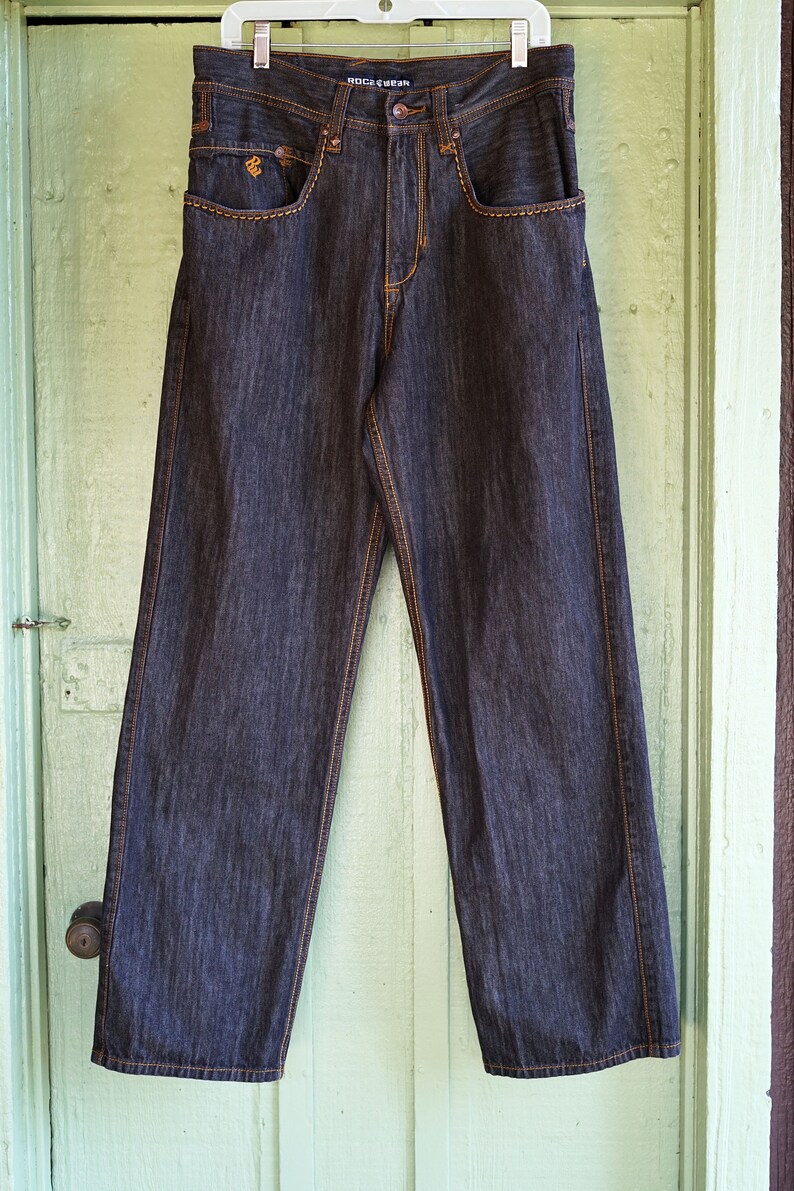 Peut inclure: Un jean en denim bleu fonc&eacute; avec des coutures orange. Le jean a une coupe d&eacute;contract&eacute;e et une jambe droite. Le nom de la marque "ROCA WEAR" est imprim&eacute; sur la ceinture arri&egrave;re.