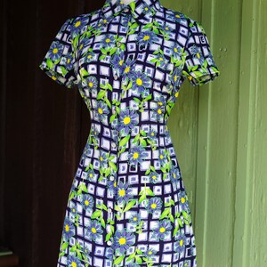 May include: A short-sleeved dress with a floral pattern. The dress features a button-down front, a collared neckline, and a fitted silhouette. The pattern includes blue flowers, green leaves, and a white grid design on a navy blue background.