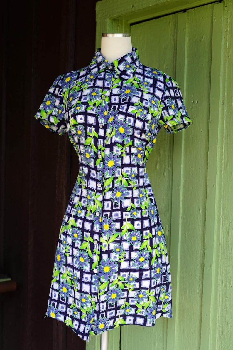 May include: A short-sleeved dress with a floral pattern. The dress features a button-down front, a collared neckline, and a fitted silhouette. The pattern includes blue flowers, green leaves, and a white grid design on a navy blue background.