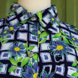 May include: Close-up of a button-down shirt with a floral pattern. The shirt features a navy blue background with a grid of white and blue squares. Blue flowers with yellow centers and green leaves are scattered across the fabric.