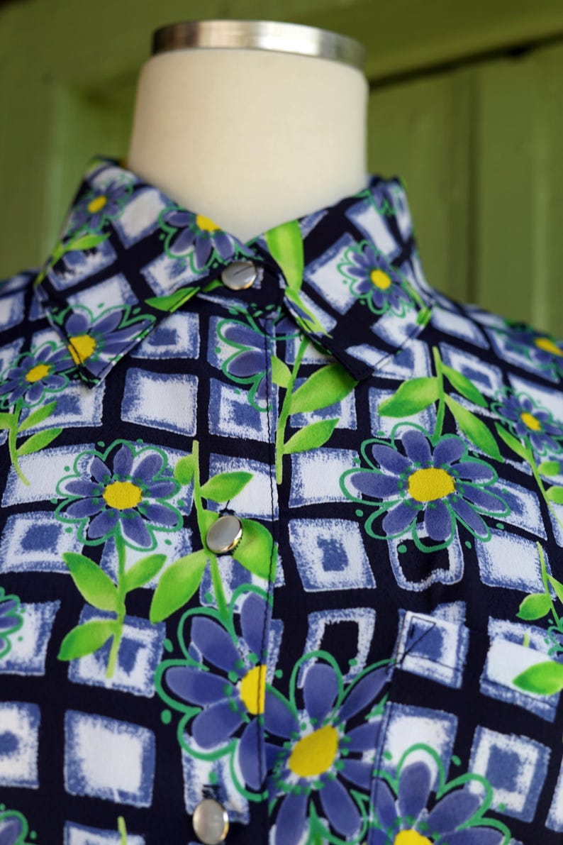 May include: Close-up of a button-down shirt with a floral pattern. The shirt features a navy blue background with a grid of white and blue squares. Blue flowers with yellow centers and green leaves are scattered across the fabric.