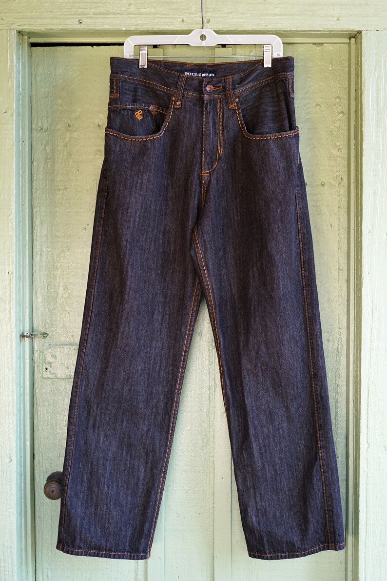 Peut inclure: Un jean en denim bleu fonc&eacute; avec des coutures orange. Le jean a une coupe d&eacute;contract&eacute;e et une jambe droite. Le nom de la marque "ROCSTAR" est imprim&eacute; sur la ceinture arri&egrave;re.