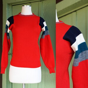 May include: Red knit sweater with white, blue, and gray striped sleeves. The sweater has a crew neck and long sleeves.