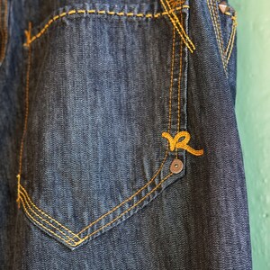 Peut inclure: Gros plan sur une poche en jean bleu fonc&eacute; avec des coutures jaunes et un petit logo brod&eacute; jaune.