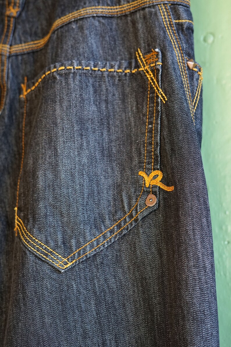 Peut inclure: Gros plan sur une poche en jean bleu fonc&eacute; avec des coutures jaunes et un petit logo brod&eacute; jaune.