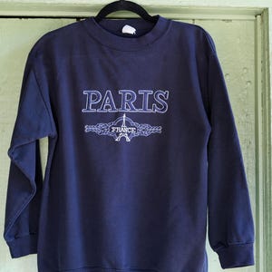 Puede incluir: Sudadera azul marino con la palabra "PARIS" bordada en blanco y azul claro, encima de un gráfico de la Torre Eiffel azul claro y la palabra "FRANCE". La sudadera está colgada de una percha negra.