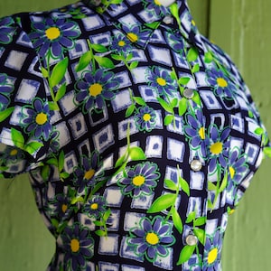May include: A vintage button-down shirt featuring a floral pattern. The shirt has a navy blue background with a grid of white squares. Blue flowers with yellow centers and green leaves are scattered across the fabric.