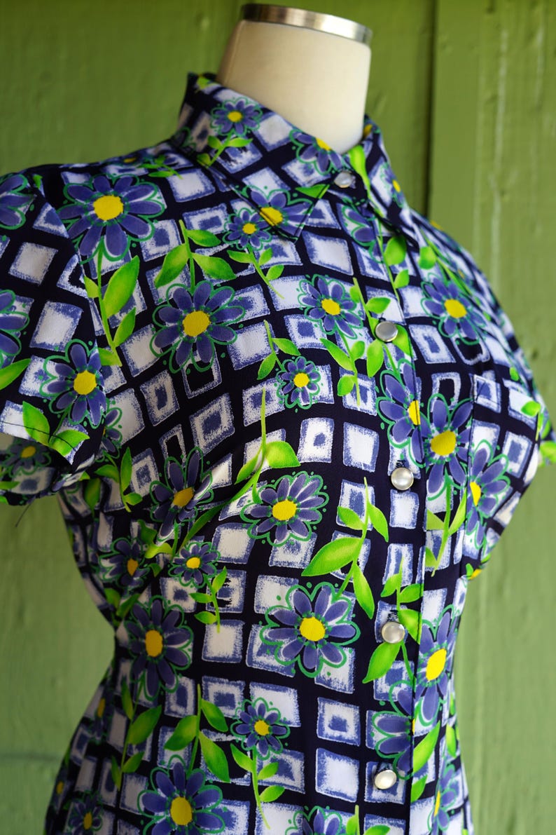 May include: A vintage button-down shirt featuring a floral pattern. The shirt has a navy blue background with a grid of white squares. Blue flowers with yellow centers and green leaves are scattered across the fabric.