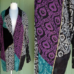 May include: A long-sleeved, multi-colored jacket with a patchwork design. The jacket is made of a variety of fabrics, including velvet, silk, and lace. The jacket has a black velvet collar and a button closure. The jacket is a mix of black, purple, teal, and silver.