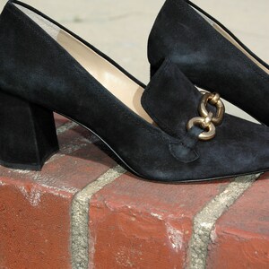 May include: Black suede heeled loafers with a gold-tone chain detail. The shoes have a square toe and a block heel. The loafers are sitting on a red brick surface.
