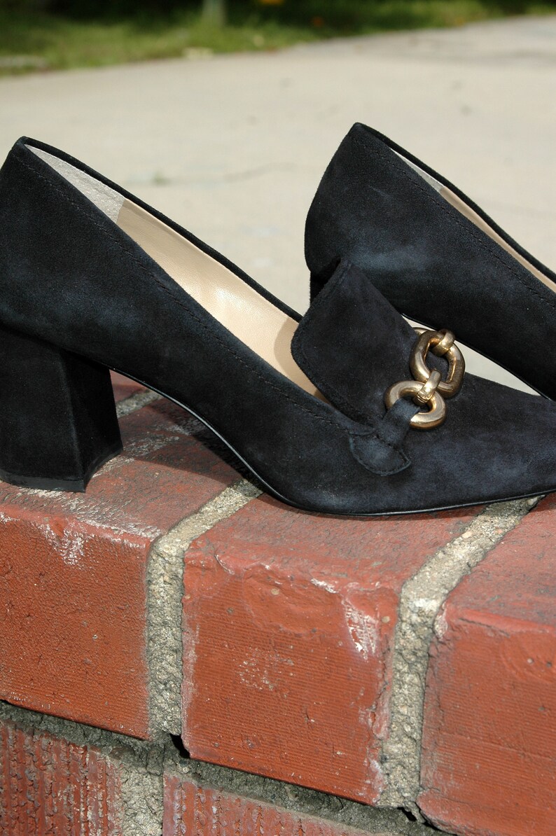 May include: Black suede heeled loafers with a gold-tone chain detail. The shoes have a square toe and a block heel. The loafers are sitting on a red brick surface.